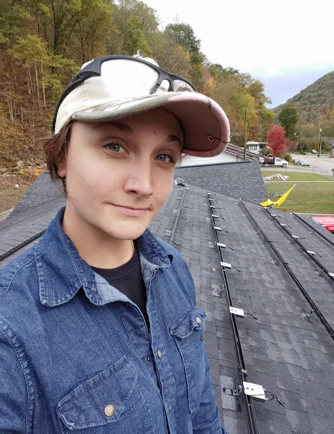 A woman with a baseball hat, protective goggles, and a collared shirt faces the camera while standing on top of a roof covered in wires and brackets to support solar panels.Trees and hills rise in the background.