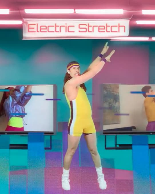 a man in a yellow unitard with purple and white stripes along the side, white socks and shoes, reaches up to the right of screen under a sign that says "Electric Stretch"