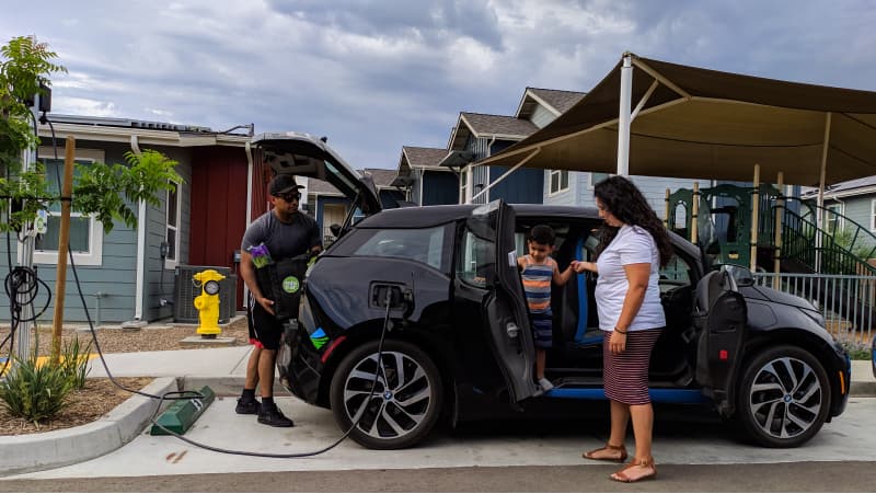 A person with long, black hair, white t-shirt and sandals stands beside the open passenger door of a black electric vehicle, holding the hand of a child in a striped peach and blue shirt inside. Another person in a hat loads groceries into the hatchback trunk, while the car is plugged in to a charger.