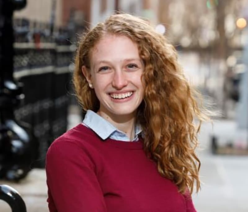 Headshot of Rebecca Federman, who is smiling and has long curly red hair. She is wearing a red long sleeve sweater with a light blue collared shirt under it.