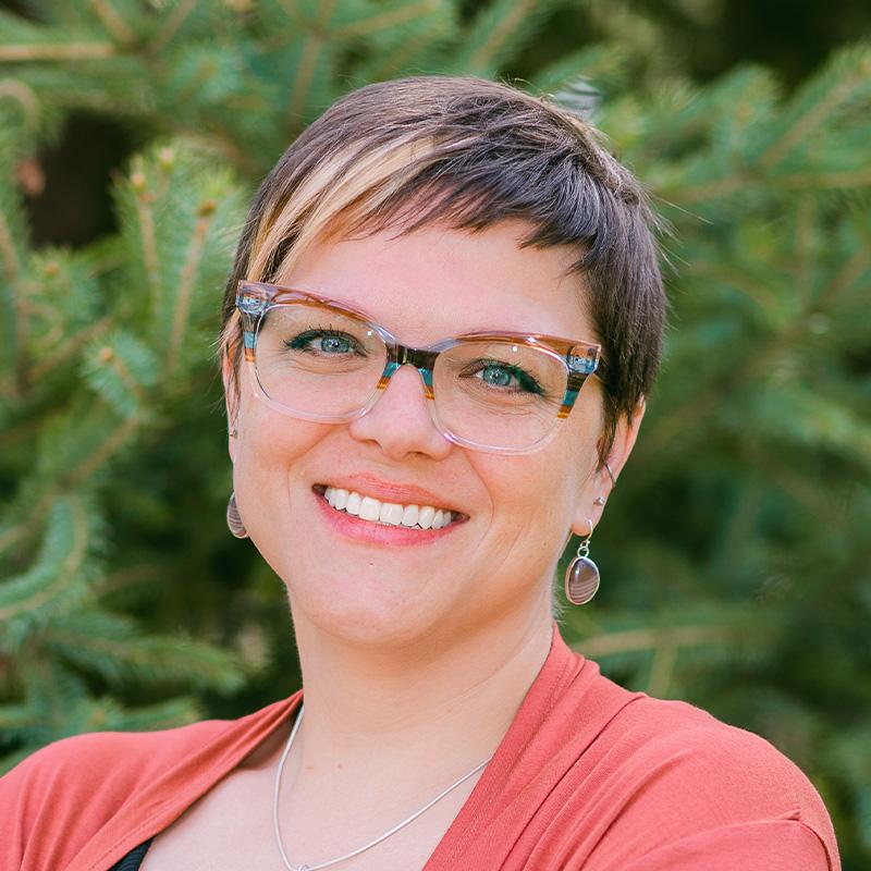 Headshot of Jenna Warmuth, who is smiling and has short light brown hair. She wears glasses and wears a salmon colored cardigan.