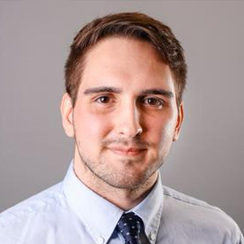 Headshot of Darren Reuter, who is slightly smiling and has short brown hair and short facial hair. He wears a light blue button up and navy tie.