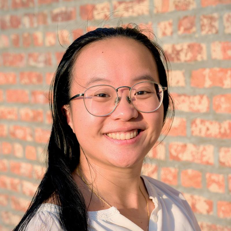 Headshot of Grace Mok, who is smiling and wears silver framed glasses. She has long black hair and stands in front of a brick wall.