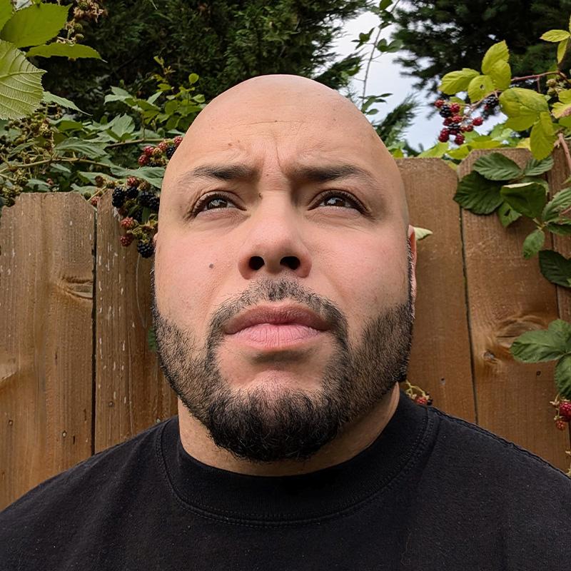 Headshot of Asaeli Matelau, who is staring into the distance. He is bald and has short black facial hair and wears a black tshirt.