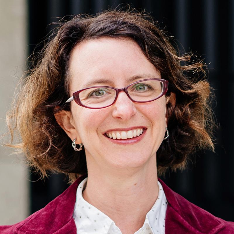 Headshot of Mary Pustejovsky, who is smiling and has chin length curly brown hair. She wears red framed glasses and is wearing a red jacket.