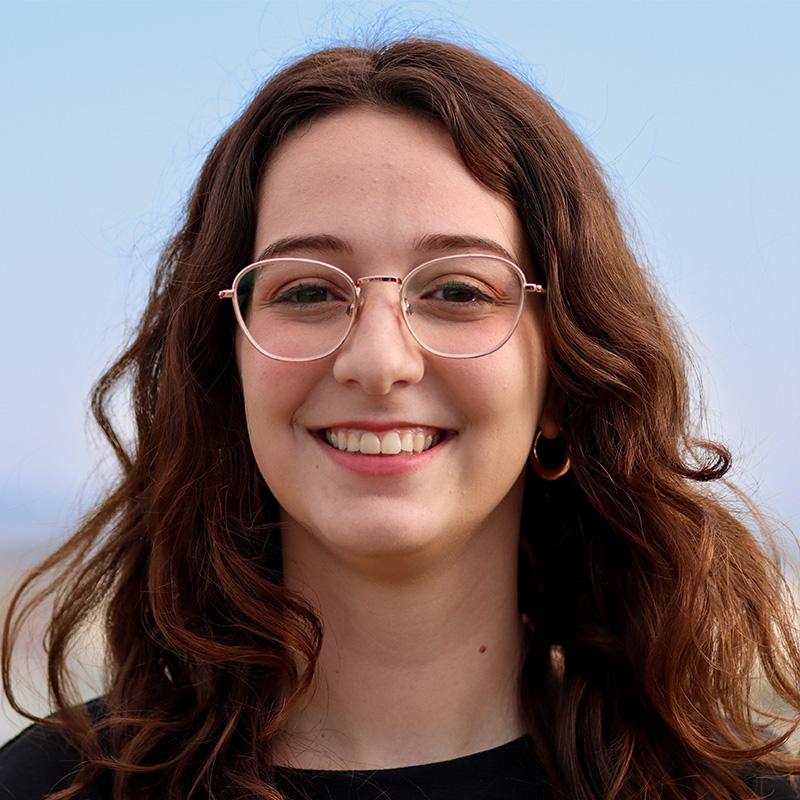 Headshot of Alana Murphy, who is smiling and has long wavy brown hair. She wears glasses and a black shirt.