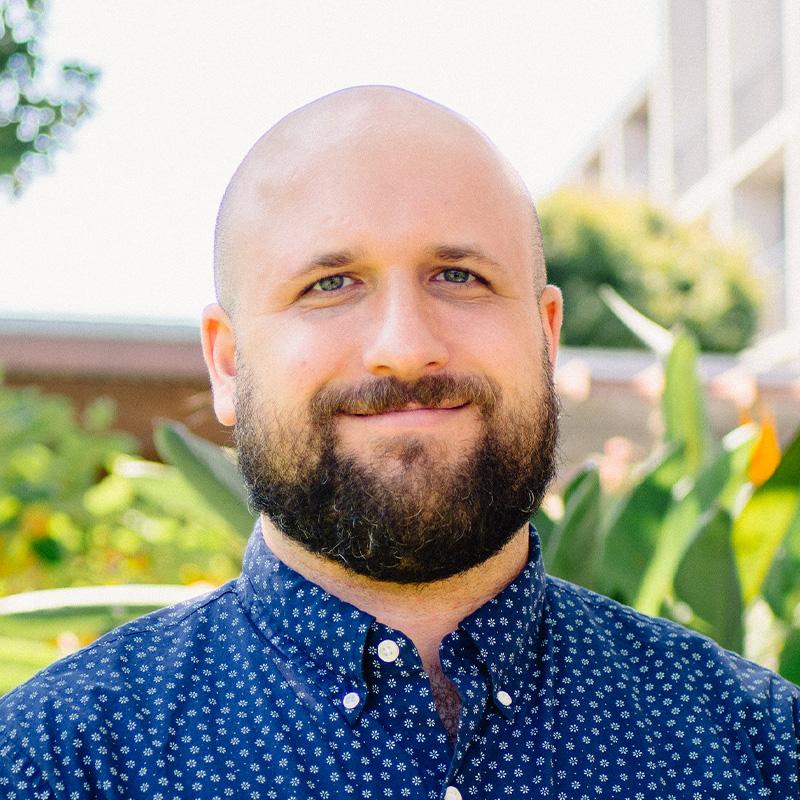 Headshot of Sam Marx, who is slightly smiling and wears a blue floral patterned button up shirt.