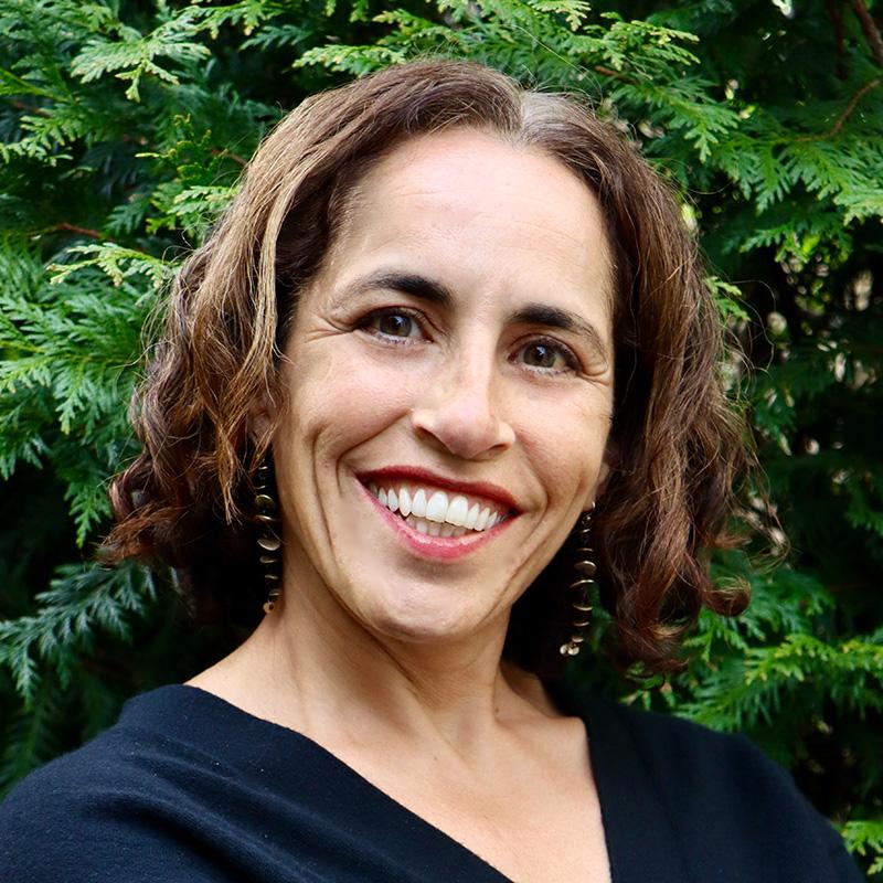 Headshot of MarjanMashhadi, who is smiling at the camera and standing in front of green trees. She has short curly light brown hair and is wearing a black shirt.