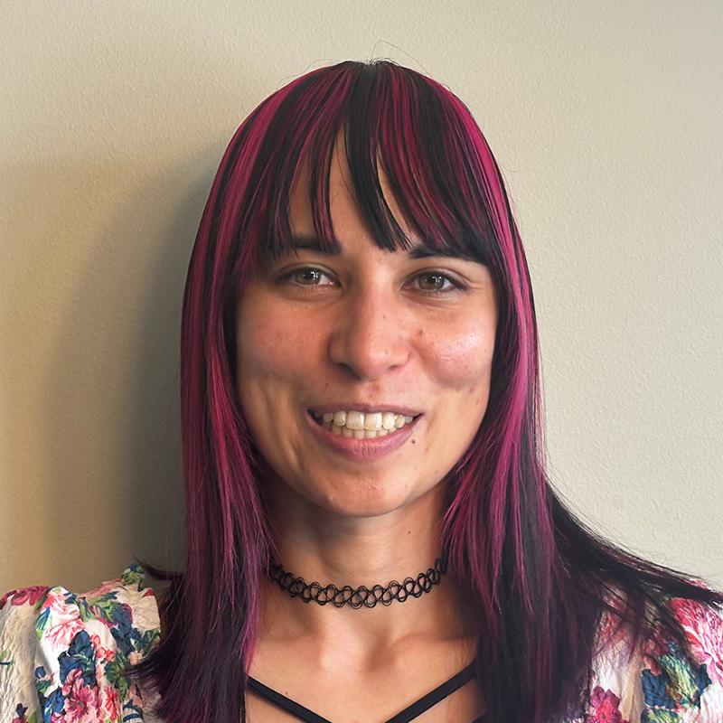 Headshot of Mindy Flores, who is smiling and has dark brown hair with bangs and pink highlights. She is wearing a floral top and a choker necklace.