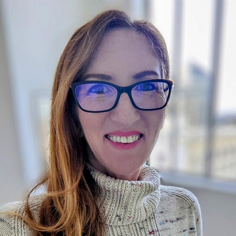 Headshot of Amy LaThanh, who is smiling and wears black framed glasses and has long reddish brown hair. She is wearing a beige turtleneck and stands in front of a window.