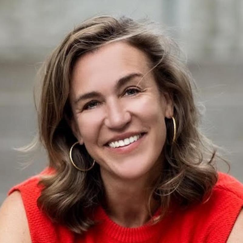 Headshot of Aimee Witteman, who smiles at the camera with her head tilted slightly to the left. She has short light brown wavy hair, wears gold hoops, and is wearing a red sleeveless top.