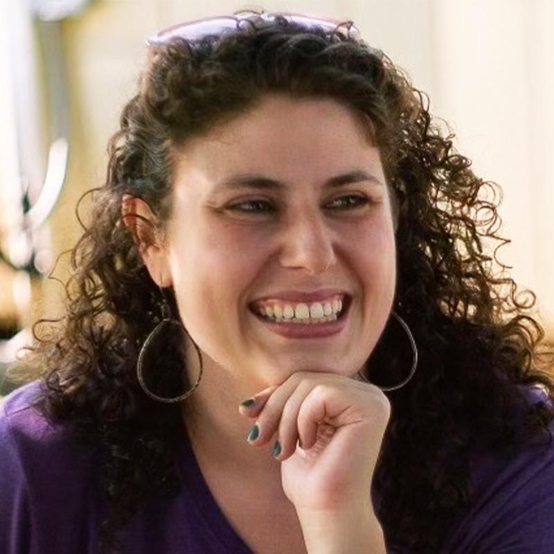 Headshot of Dorie Klein, who is smiling and looking to the right. She has curly brown hair and is wearing hoops.