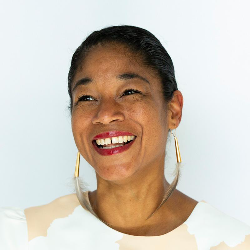 Headshot of Natalie Bomberry, who is smiling and has black hair pulled back into a ponytail. She wears feather earrings, red lipstick, and a beige top.