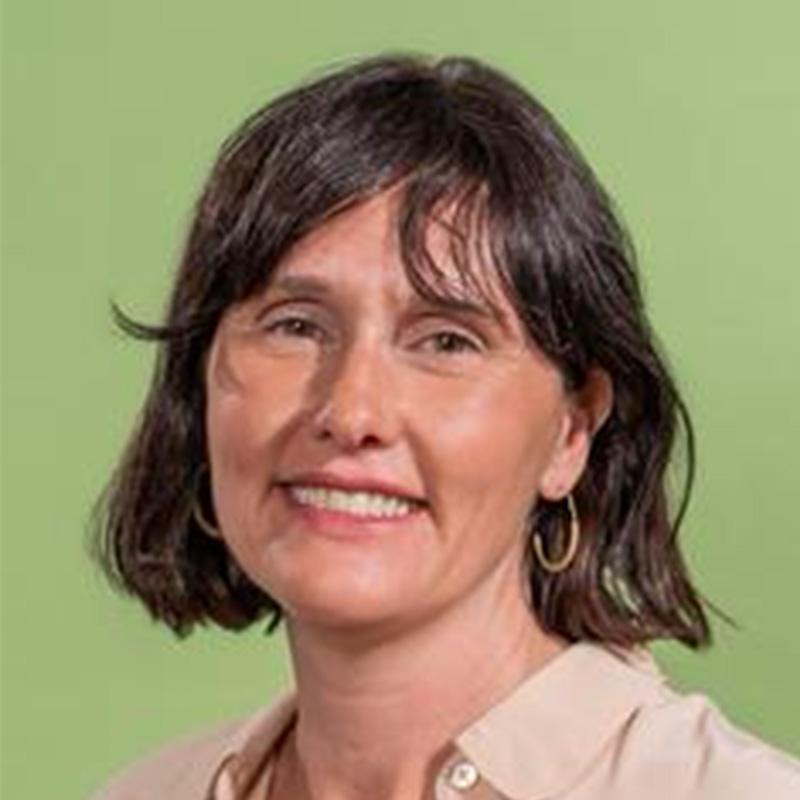 Headshot of Tracey Patterson, who is smiling and has short brown hair and bangs. She is wearing a beige top and stands in front of a green background.