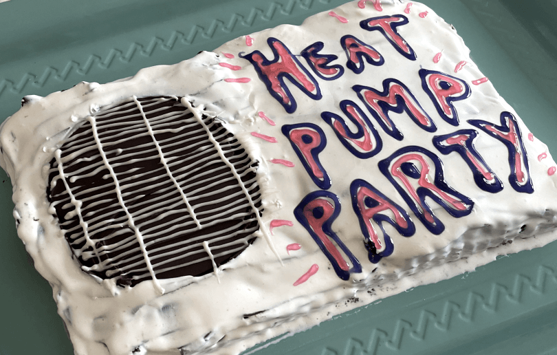 Homemade cake with white frosting and pink icing that says 'Heat pump party'. The cake is decorated to look like a heat pump and is placed on a greenish-blue tray.