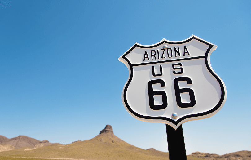 White Arizona US route 66 sign in front of a clear blue sky with rock formations in the background.