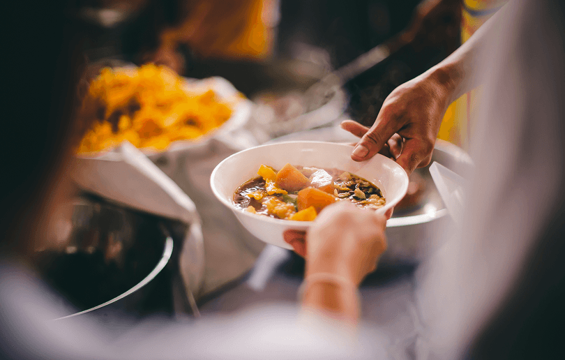 A person's hand passes a steaming bowl of soup to another person. The soup has large chunks of sweet potato and other vegetables inside it.