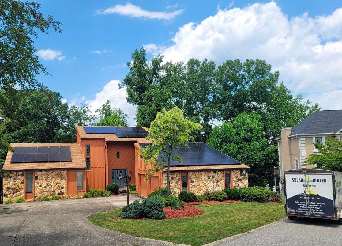 A concrete driveway leads up to a a split-level home with solar panels covering the roof. The home is surrounded by trees and a bright, sunny sky. A truck with the name "Solar Holler" is parked alongside.